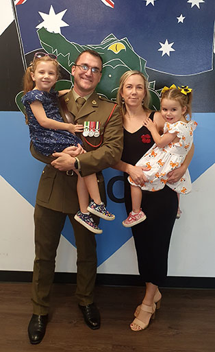 An army officer in full uniform holding his daughter in his arms. Next to him a woman with another daughter.